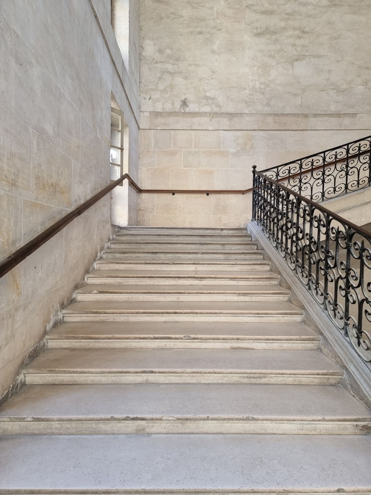 Grand escalier de pierre à volées droites et rampe en fer forgé, dans un immeuble ancien.