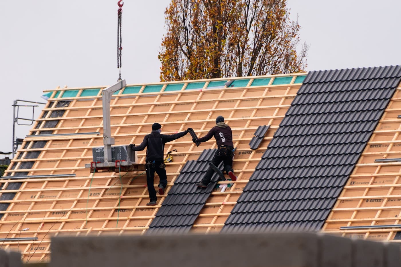 Deux couvreurs, vus de dos, posant des tuiles mécaniques sombres sur une grande toiture neuve : chevrons et liteaux clairs, écran de sous-toiture et monte-matériaux en pied de versant.