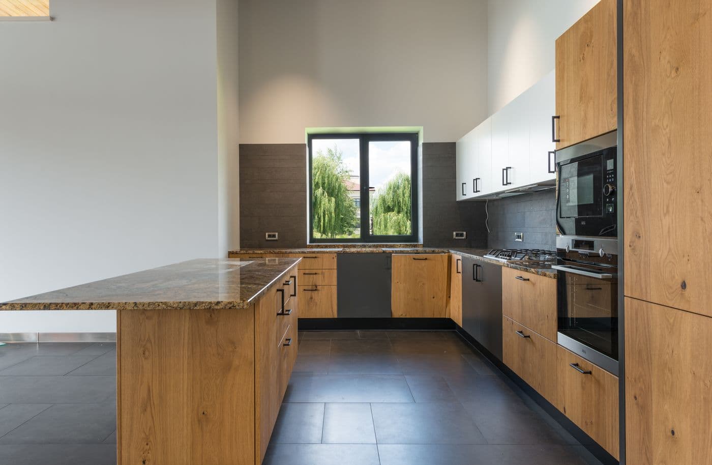 Cuisine finie à façades en bois clair, plan de travail en granit et crédence carrelée près d'une fenêtre.