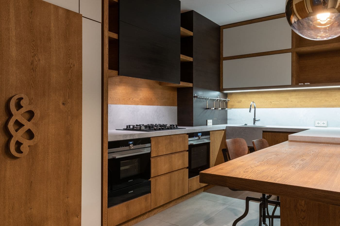 Cuisine contemporaine à façades en chêne et éléments noirs : plaque gaz, fours encastrés et table-bar en bois.