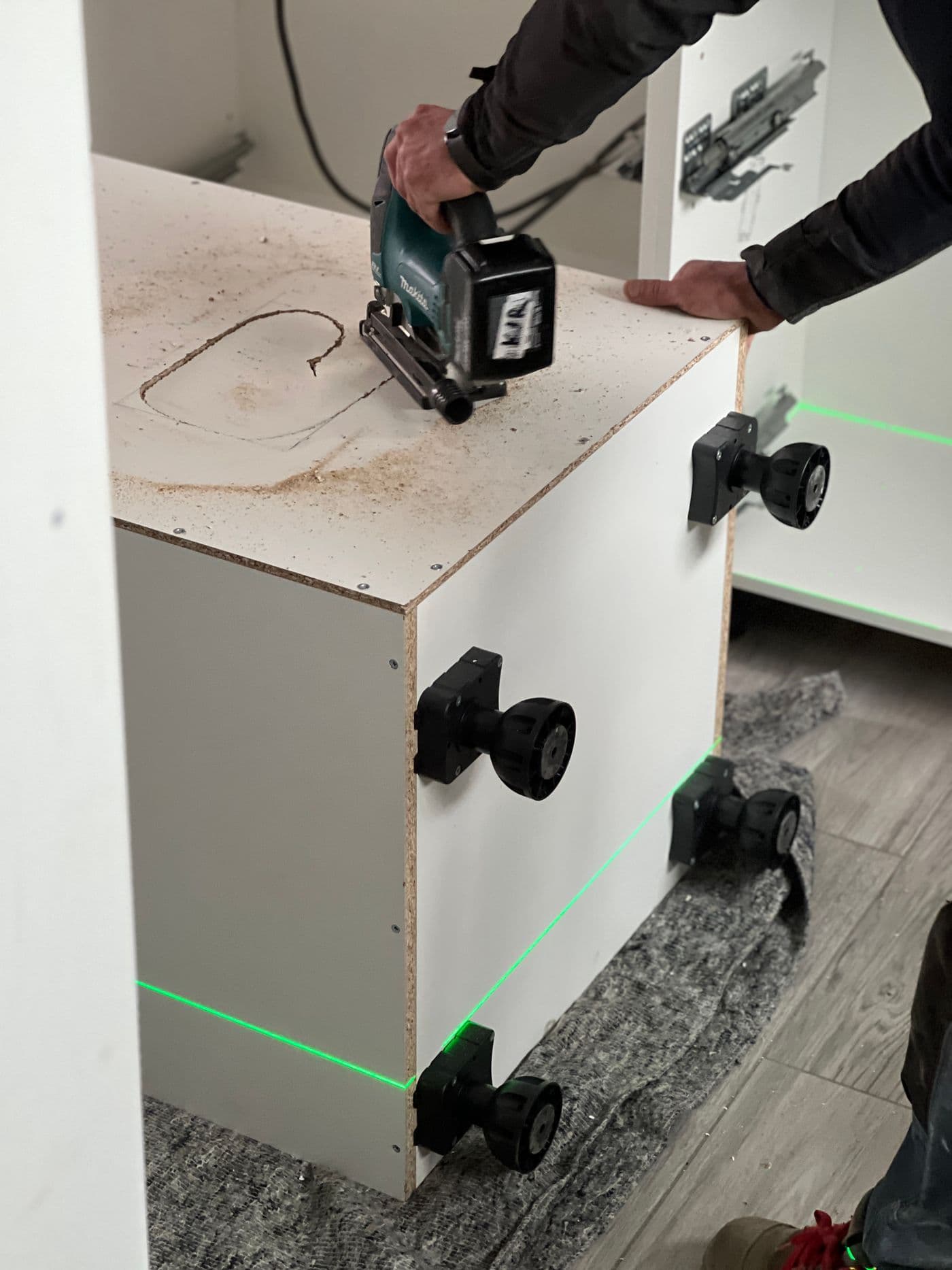 Pose d'une cuisine : découpe d'un caisson à la scie sauteuse, pieds réglables et niveau laser sur le sol.