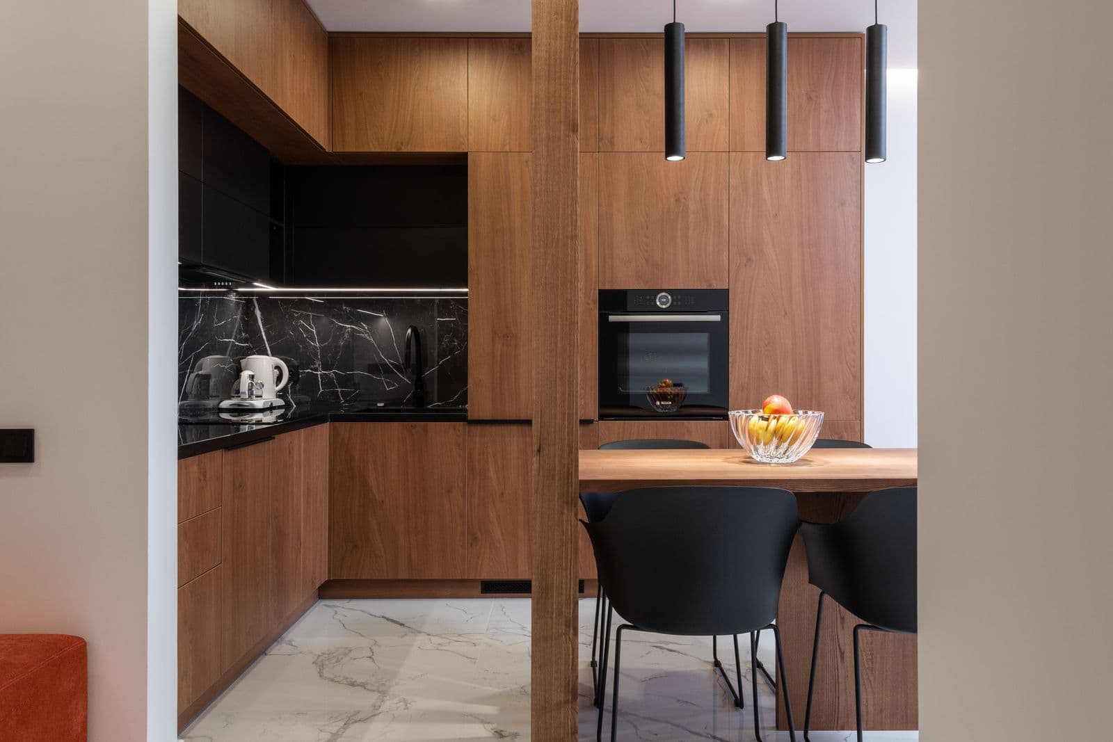 Cuisine en noyer à plan noir, coin repas avec table en bois et suspensions, sol en marbre clair.