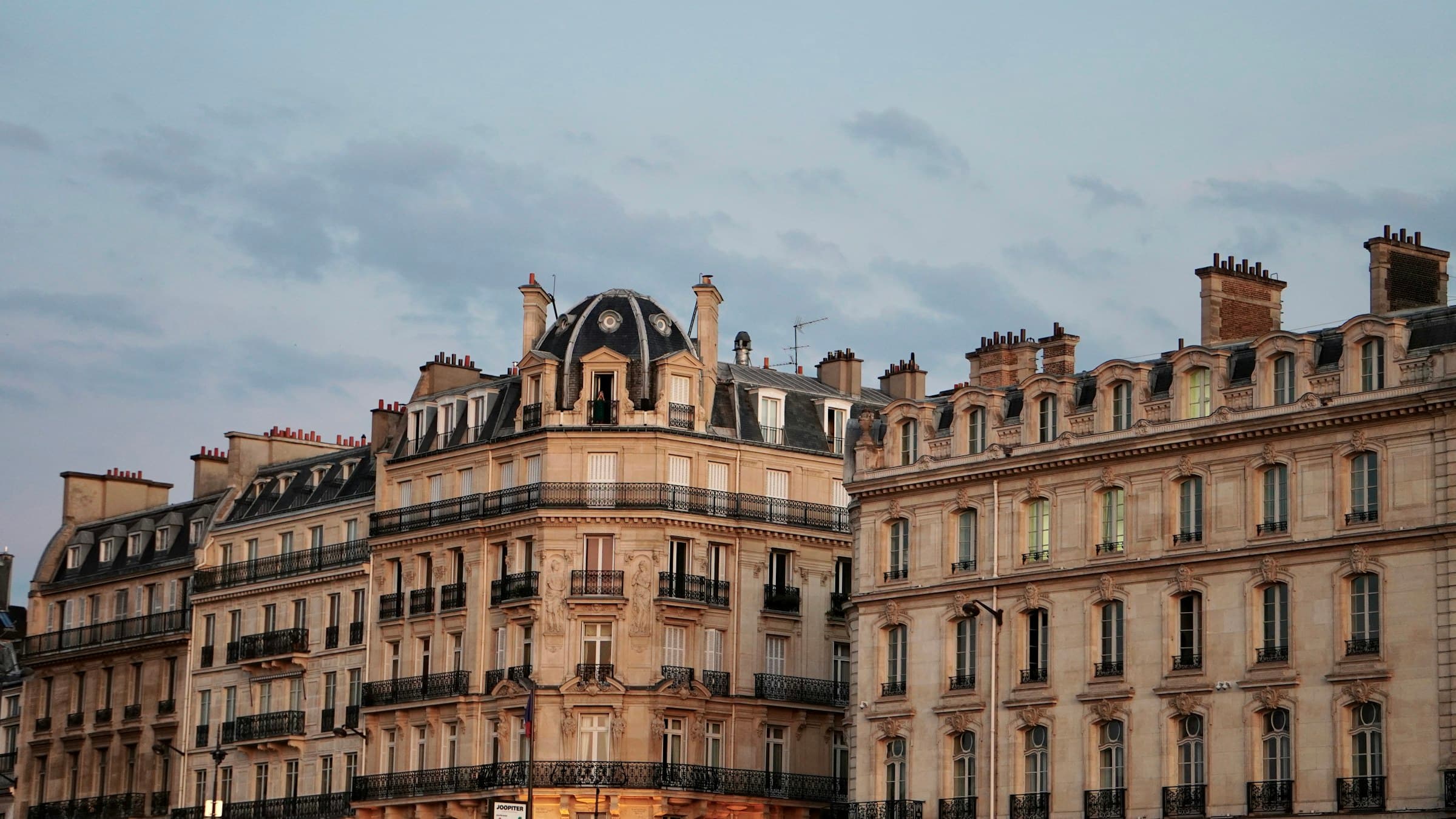 Immeuble haussmannien d'angle au crépuscule : façade en pierre de taille, dôme en ardoise et balcons filants en fer forgé.