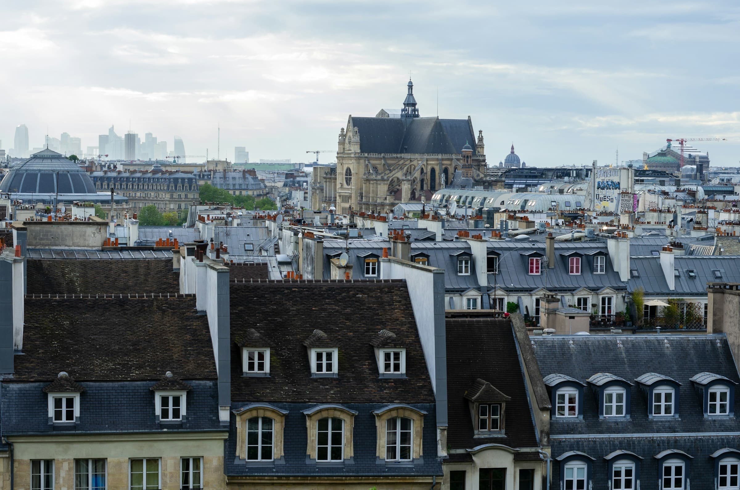 Vue en surplomb des toits de Paris : toitures en zinc et en ardoise, lucarnes à fronton et souches de cheminée coiffées de mitrons en terre cuite.