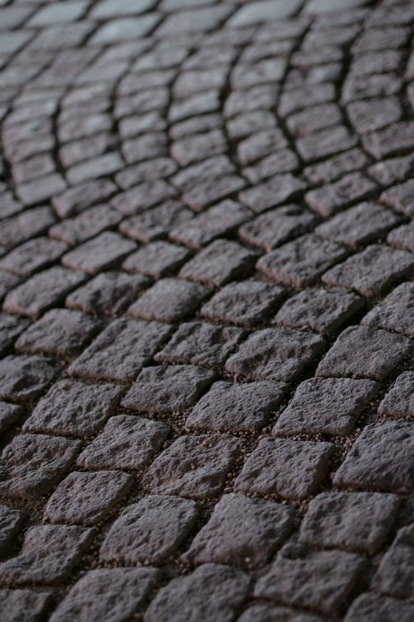 Pavage ancien en pavés de pierre posés en éventail (queue de paon), joints serrés et tons chauds sombres.