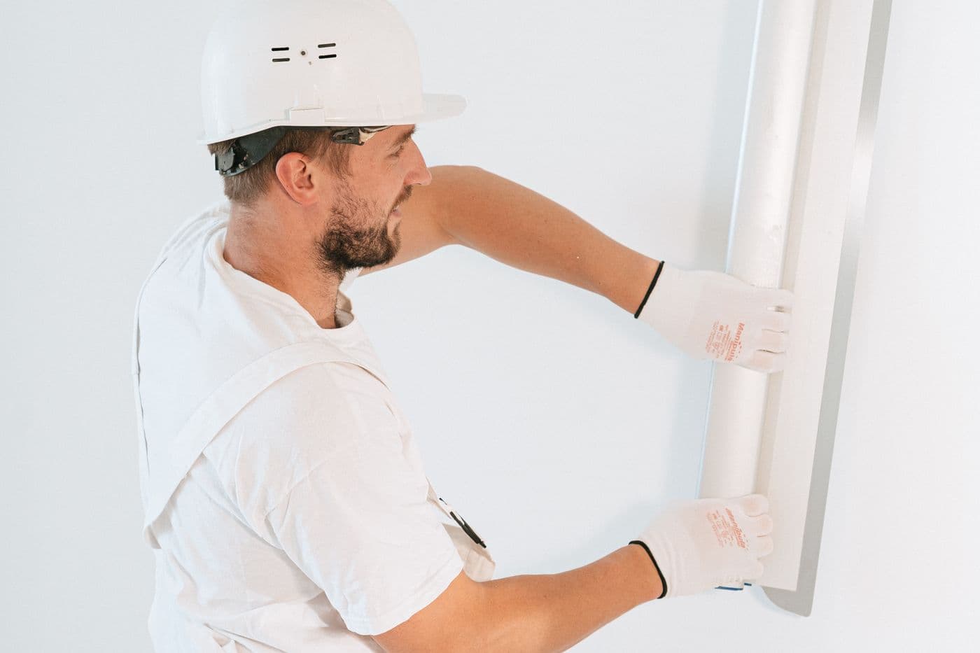 Ouvrier en casque et gants posant une plaque de plâtre contre un mur, sur un chantier de cloison sèche en rénovation.